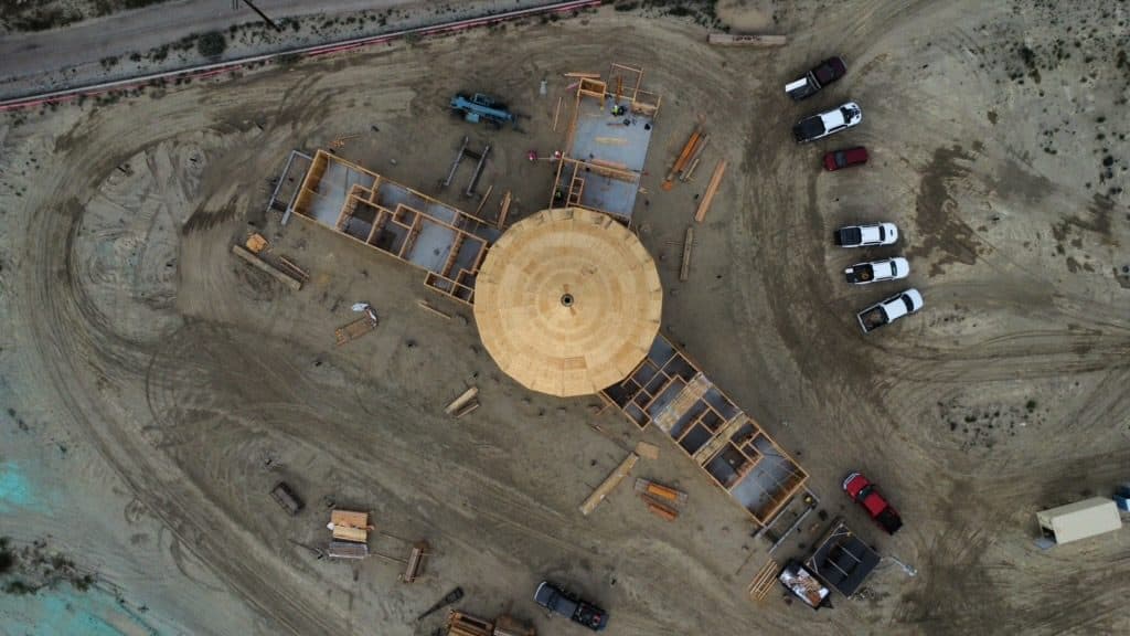Straight-down aerial of the great room's circular plywood deck, lumber stacked beside it on the pad and pickups parked at the edge.