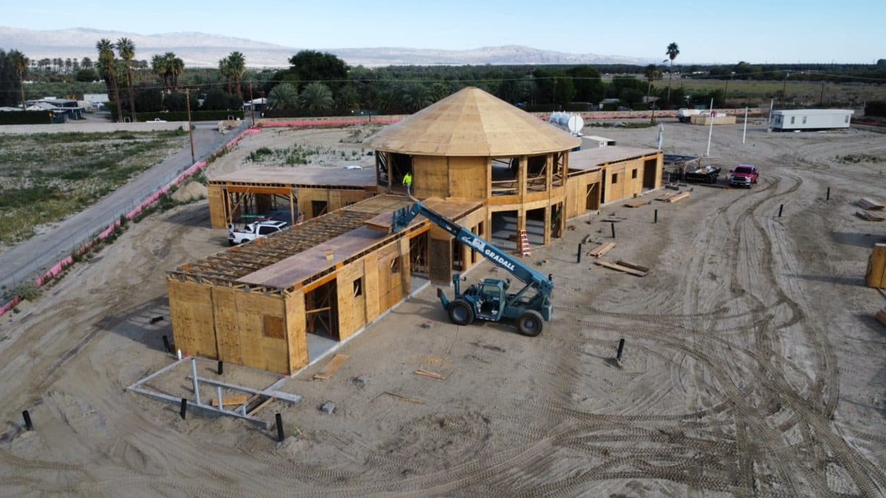 Low aerial of the estate in framing: the two-story round center mass rising first, a telehandler working beside it, and date palms and farmland stretching beyond the pad.