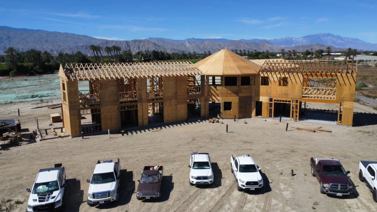 Aerial front view of the fully framed estate, a row of crew trucks parked along the dirt frontage and desert mountains across the valley behind.