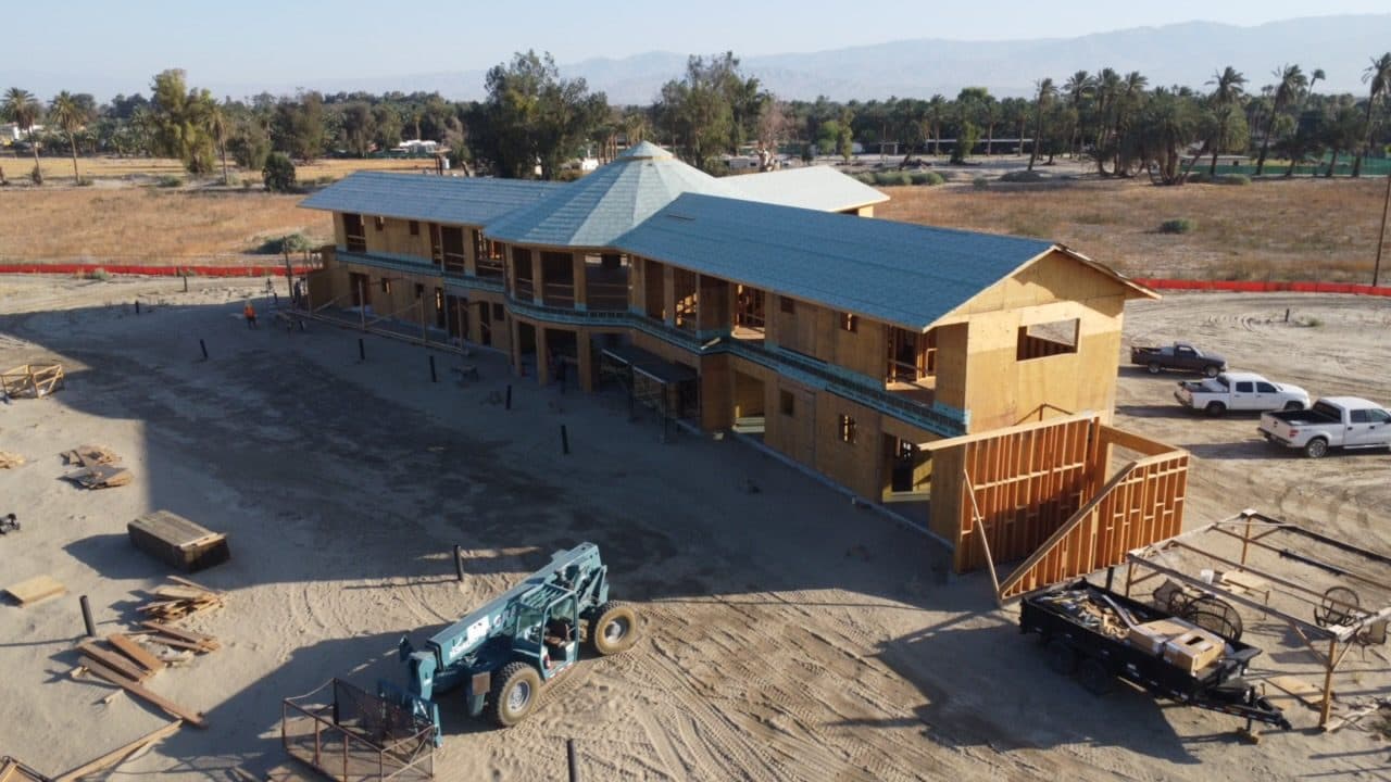 Aerial of the roofed estate from the rear: green roofing over both wings, a second-floor walkway running the length of the elevation, and a telehandler staged below.
