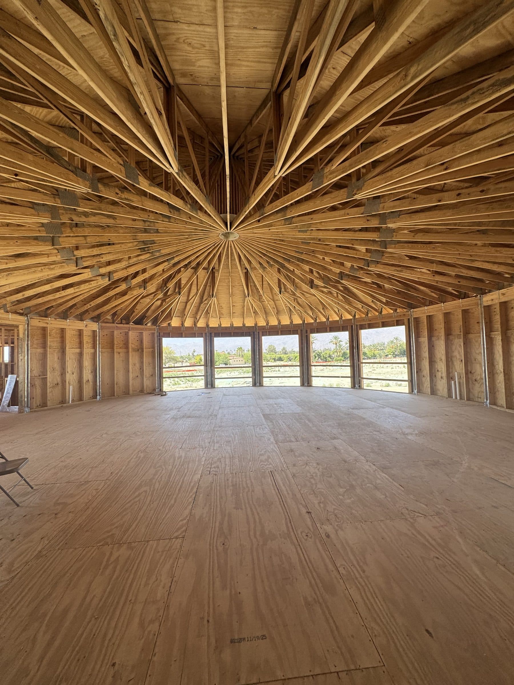 Inside the completed frame of the great room: dozens of radial rafters meeting at the steel hub, window openings following the curve of the wall, green fields beyond.