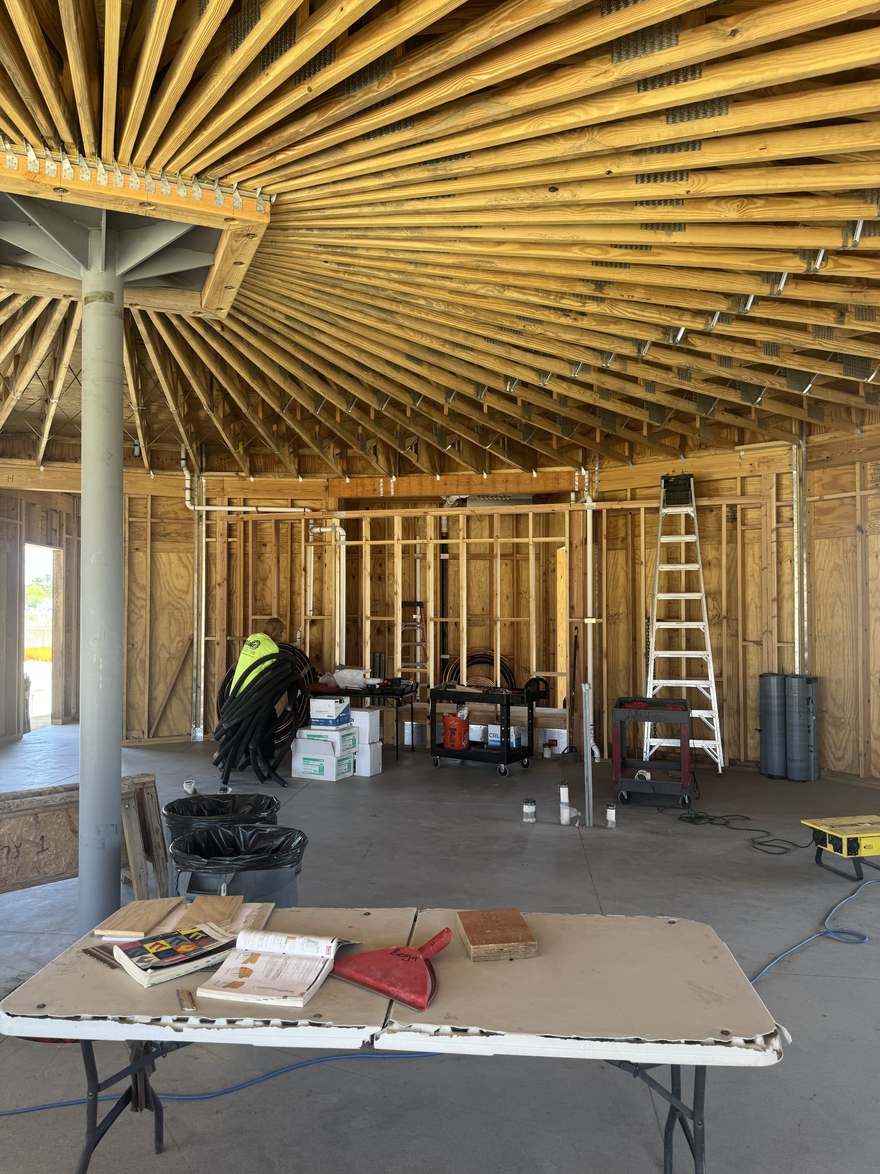 A steel column carrying a radial fan of framing overhead, with a folding work table and jobsite gear on the concrete slab below.