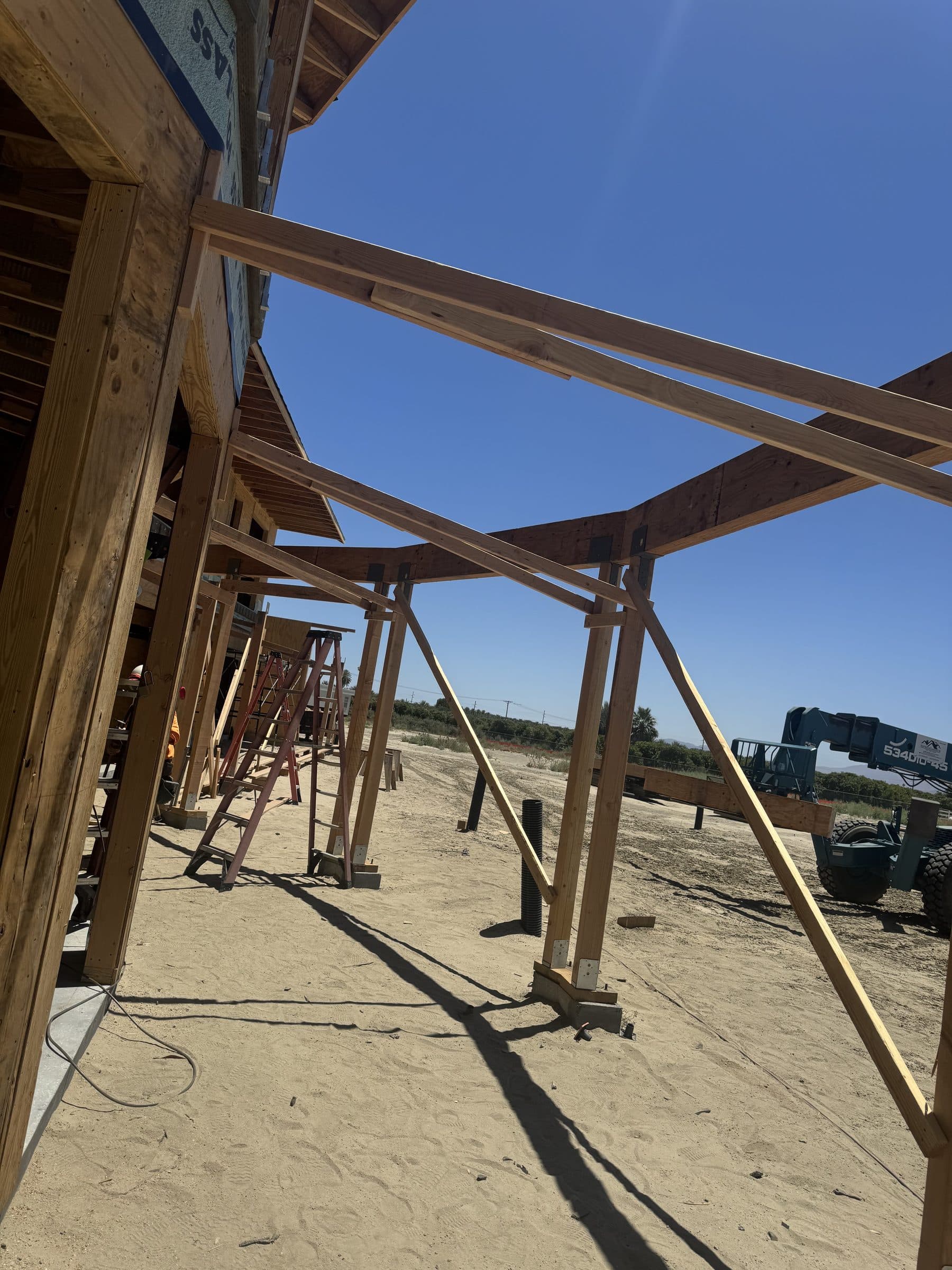Heavy timber posts and beams of a covered walkway in framing, running across the open pad under a clear desert sky.