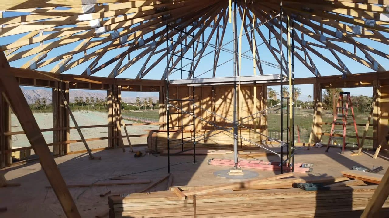 Inside the round room during roof framing: bare radial rafters overhead, a steel center column inside a scaffold tower at the middle of the deck, and the green valley floor beyond.