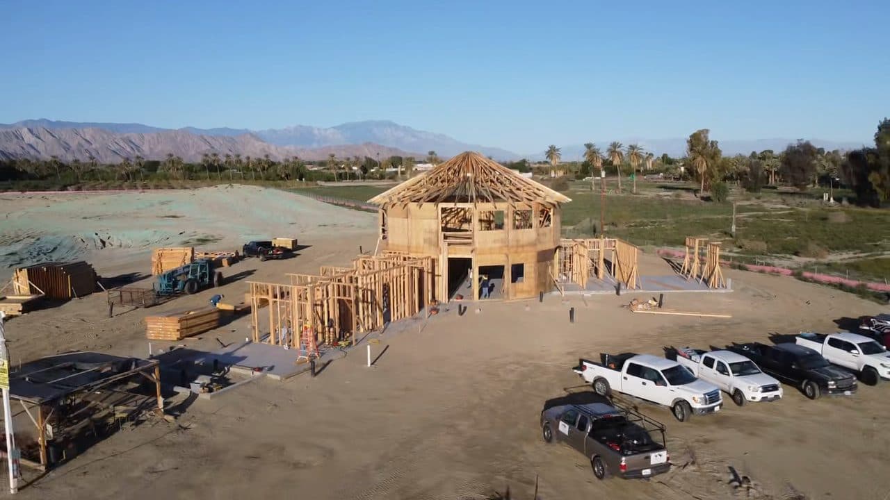 Low aerial during framing: the two-story round center rising above first-floor walls, pickups along the road edge, mountains across the valley.