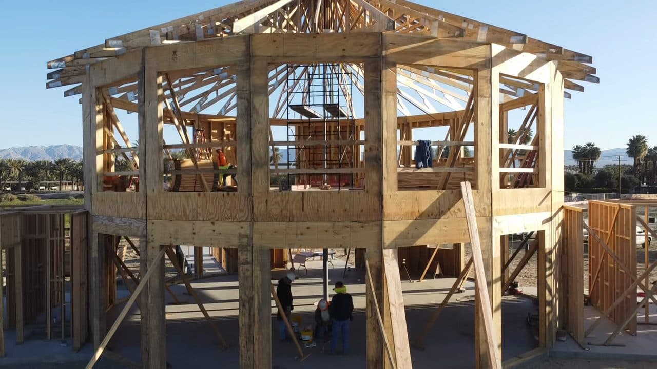 Framing crew at work inside the two-story drum of the great room: braced posts and ring beams around them, roof spokes overhead, and a work tower rising through the center.