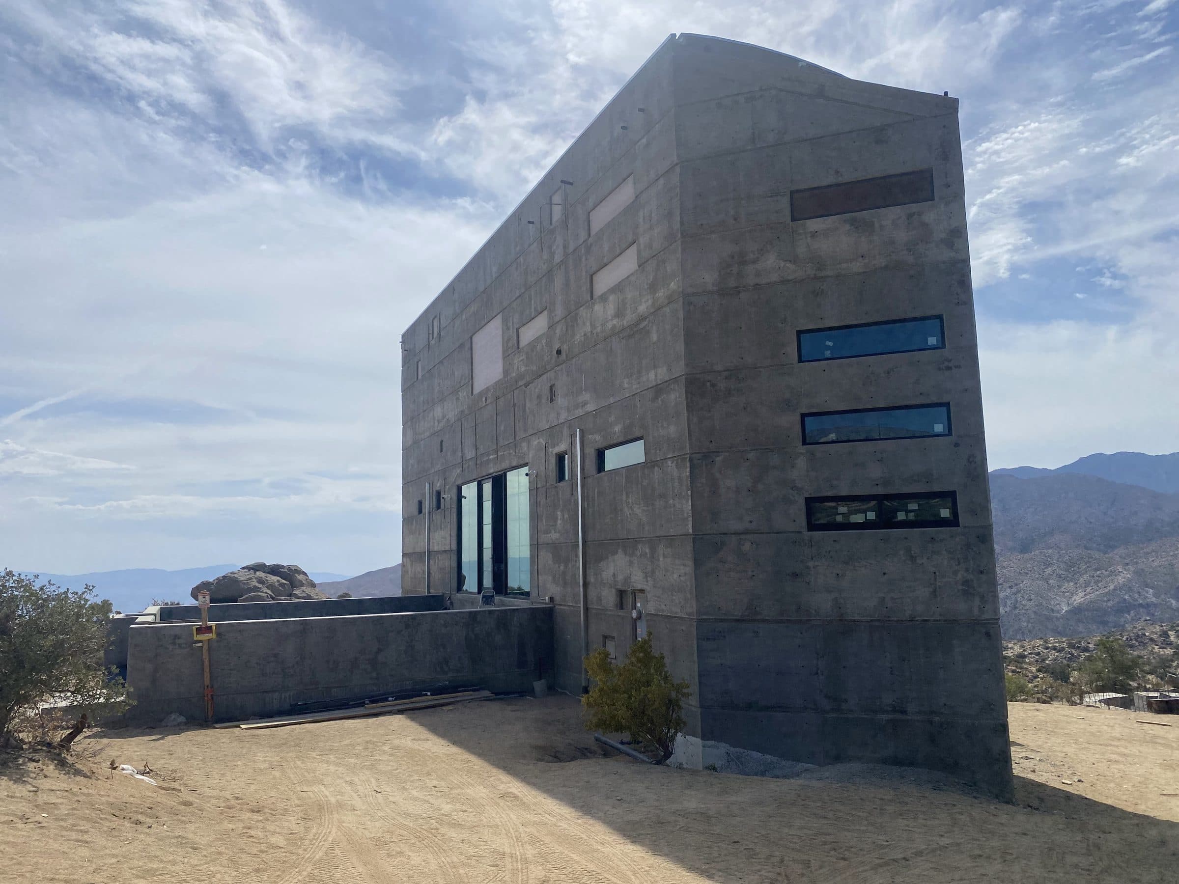 Board-formed concrete house on a desert ridge, slot windows stepping across its banded face, hazy ranges beyond.