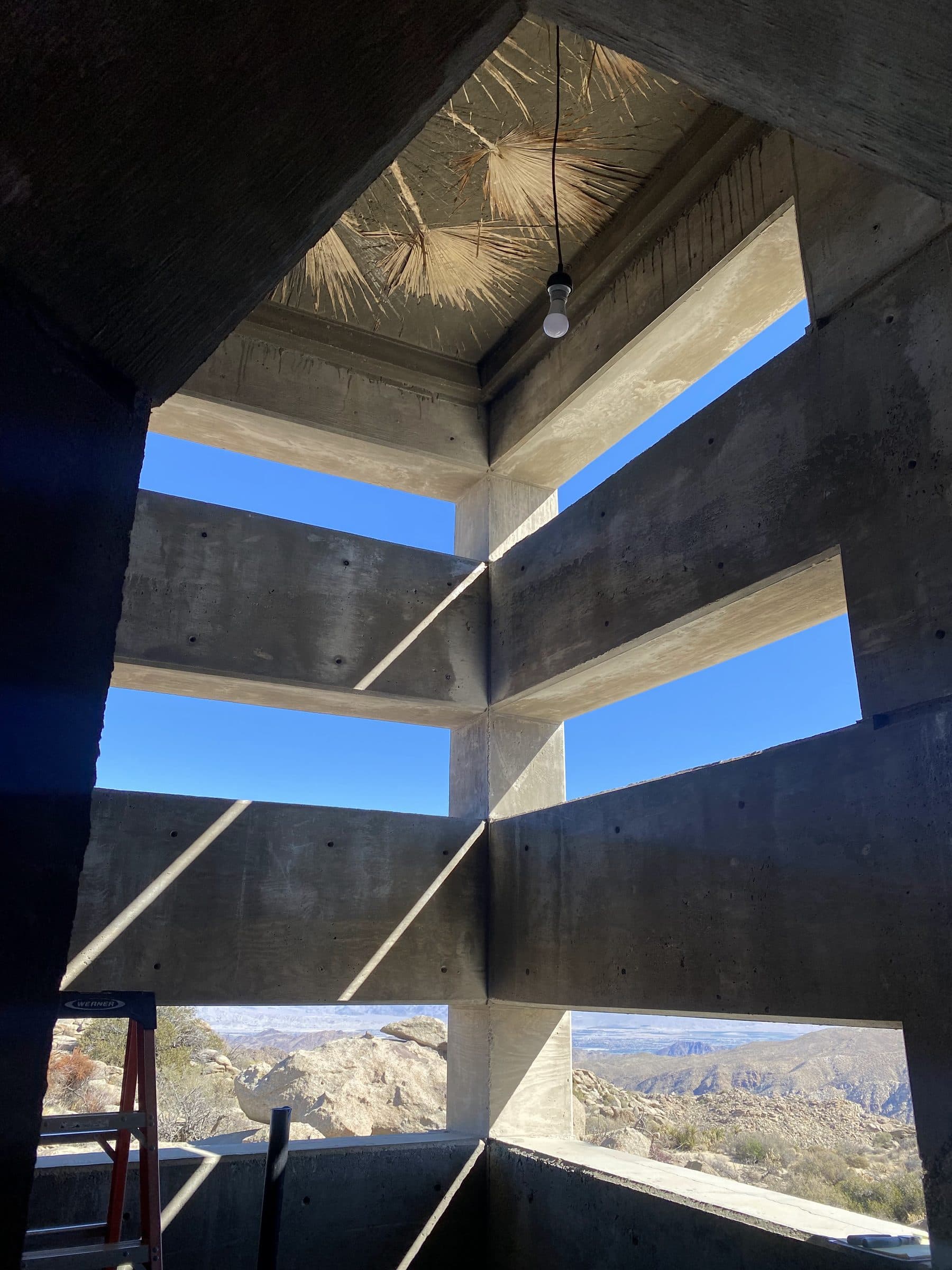 Corner of stacked concrete window slots from inside, a bare bulb hanging from a palm-frond ceiling, desert valley beyond.