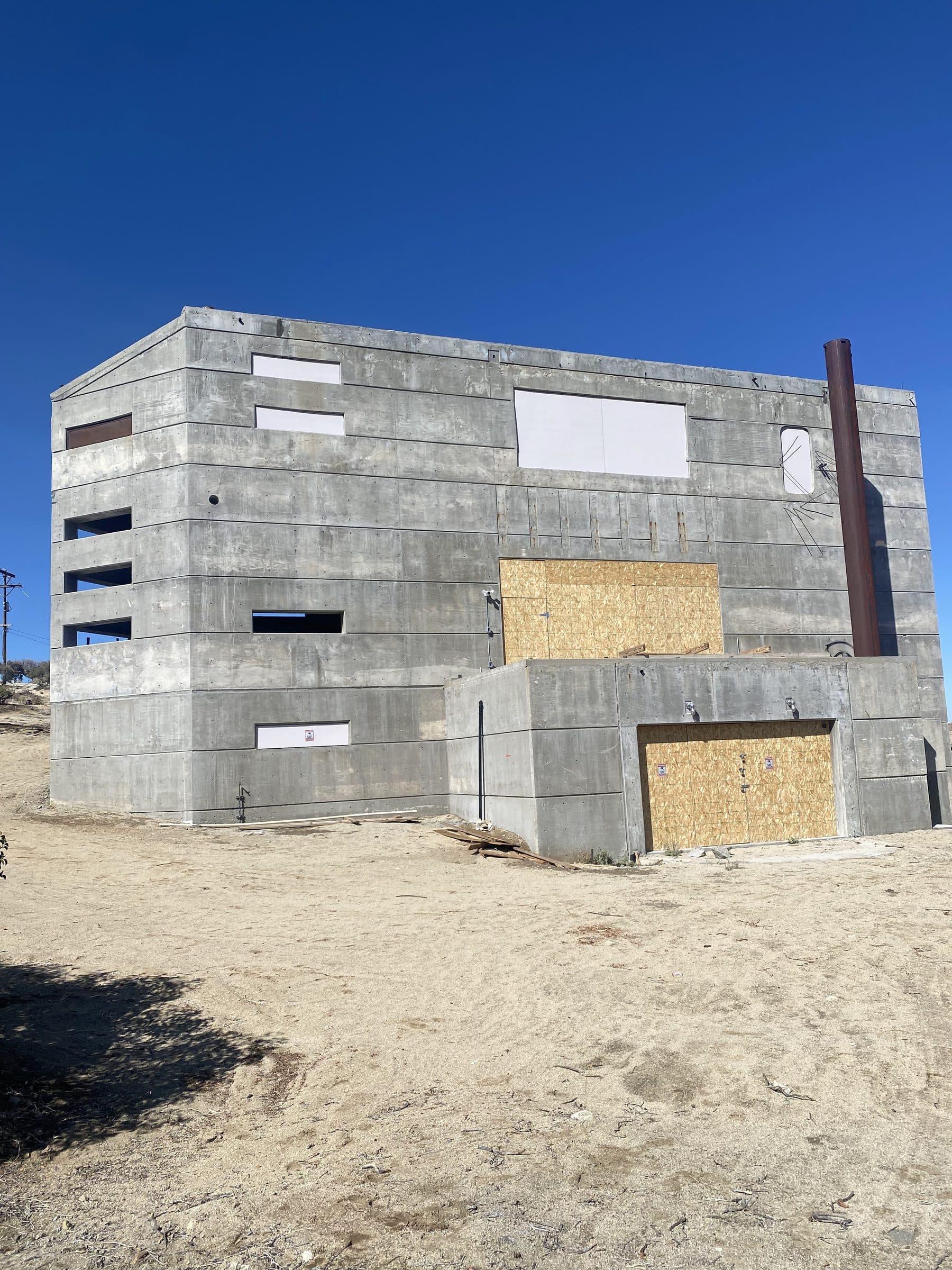 Weathered board-formed concrete shell with boarded openings, a rusted steel flue on its right face, deep blue sky.
