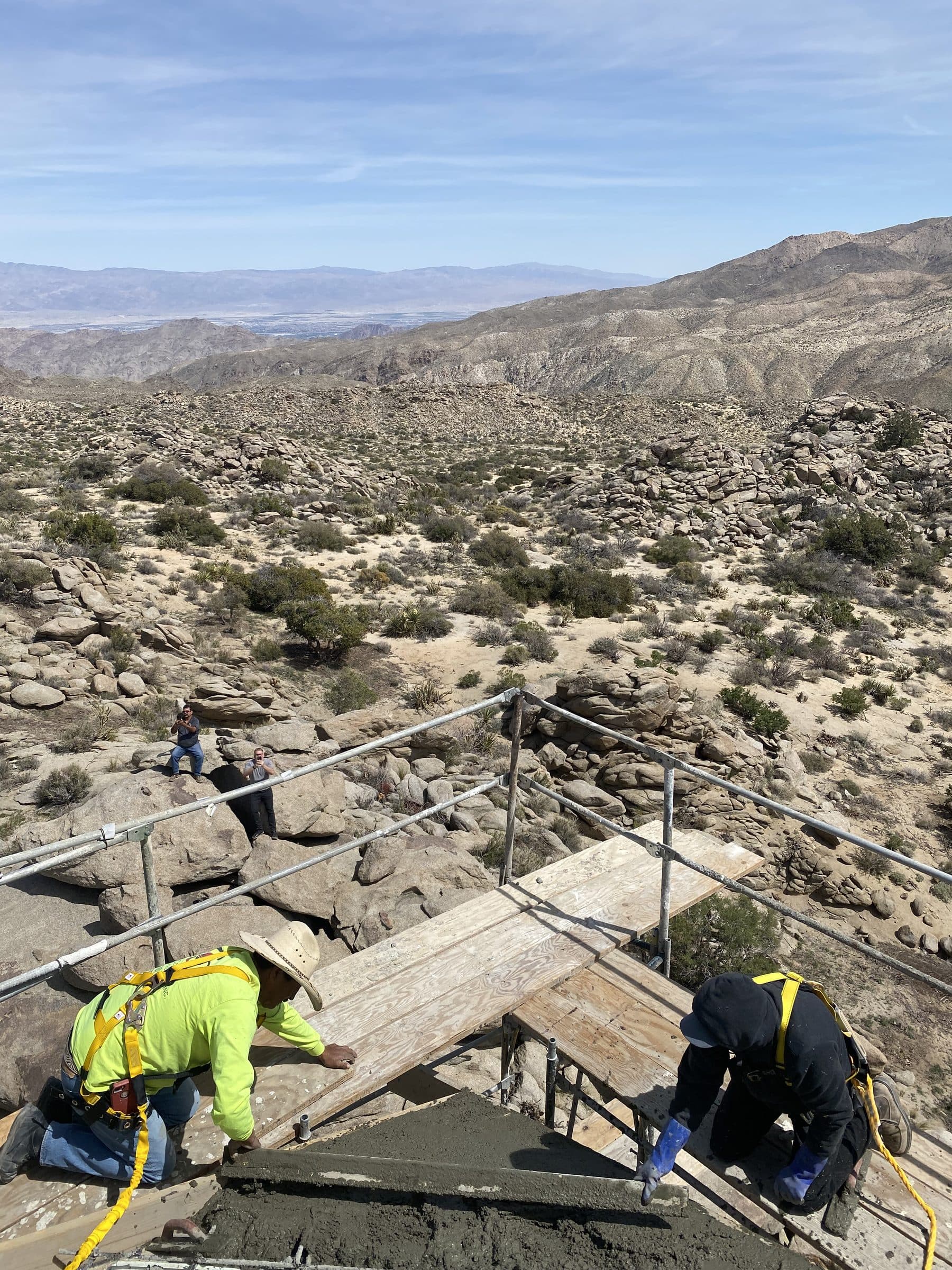 Crew screeding wet concrete at the scaffolded roof edge, boulder field and the desert basin far below.