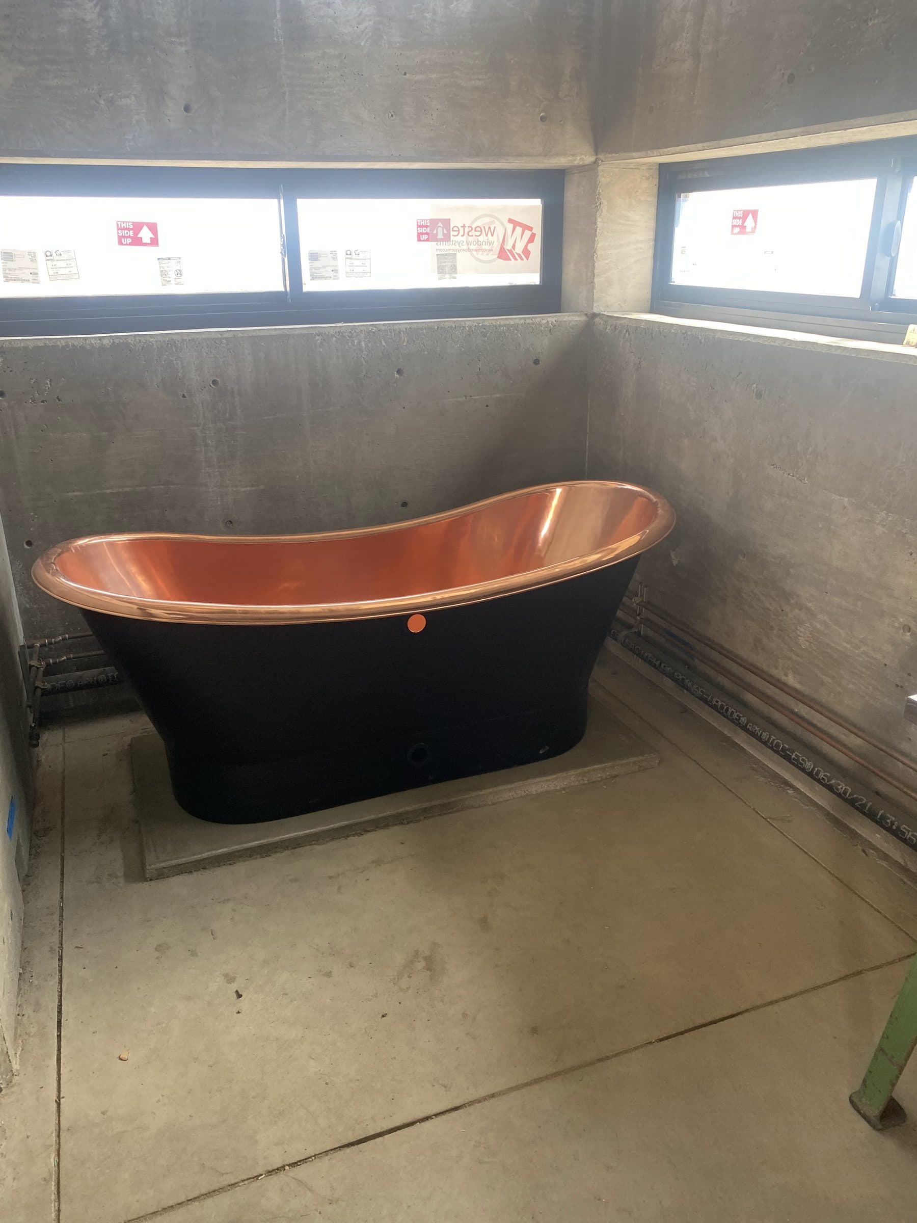 Black slipper tub with a polished copper interior on a bare concrete floor, ribbon windows above.