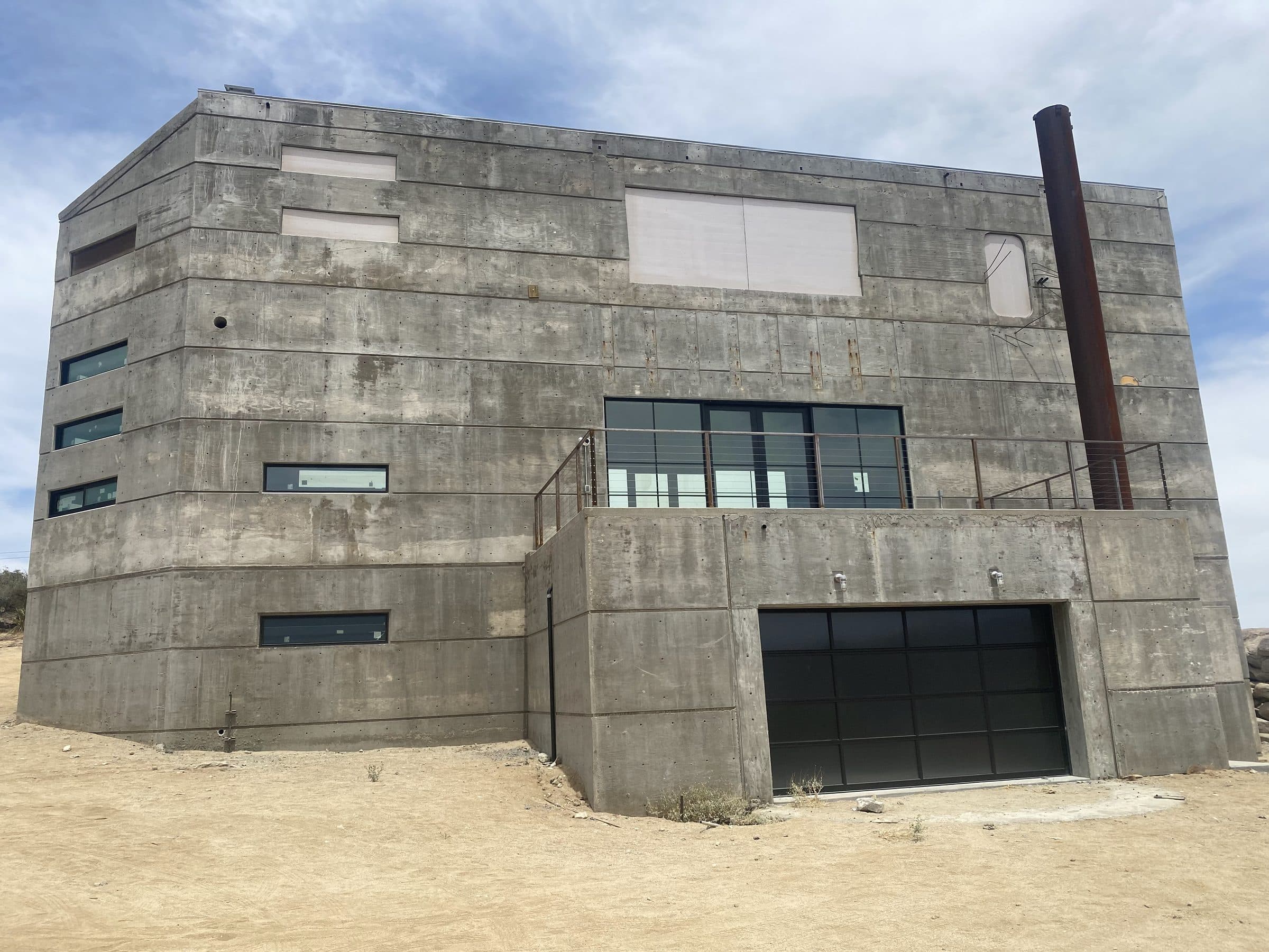 Finished concrete shell in afternoon light: banded board-formed walls, garage volume at right, rusted steel flue against the sky.