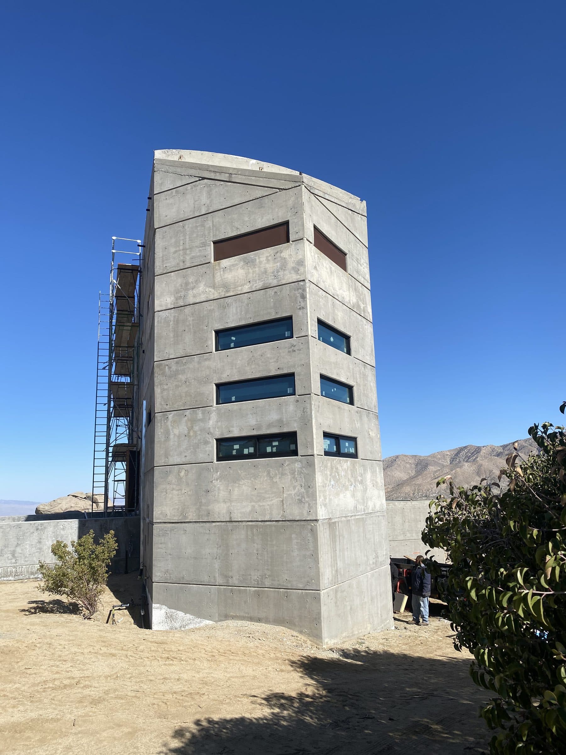 Faceted concrete prow with glazed ribbon windows, scaffolding at its left face, deep blue sky.