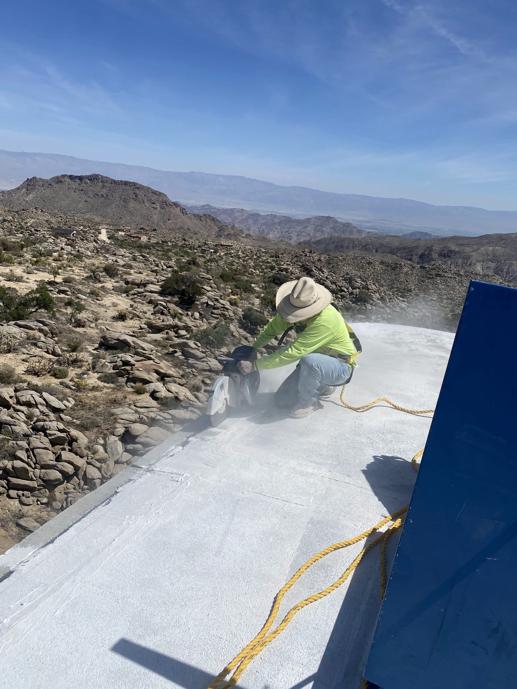 Worker in a straw hat saw-cutting the white-coated roof edge, dust drifting, the valley floor far below.