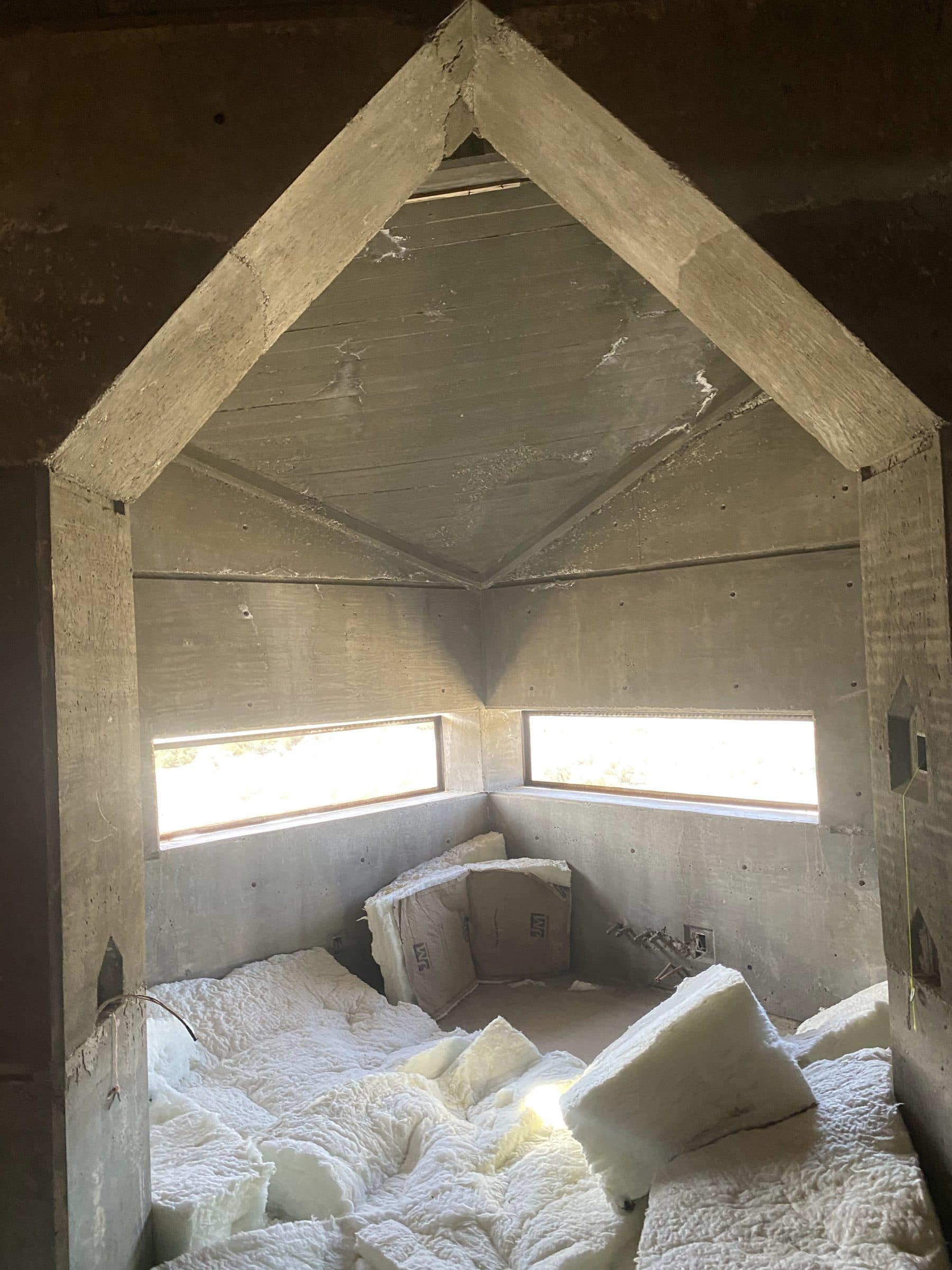 Peaked cast-concrete room with paired angled clerestory slots, insulation staged on the floor.