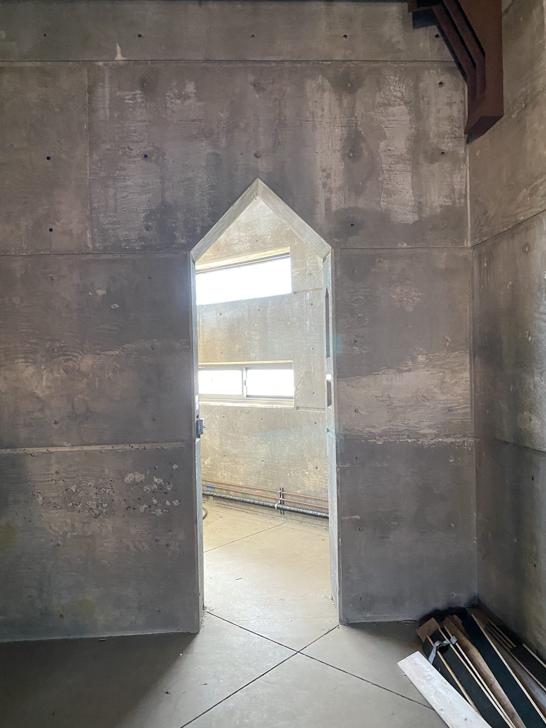 Pointed doorway cast in a board-formed concrete wall, lit window bands in the room beyond.
