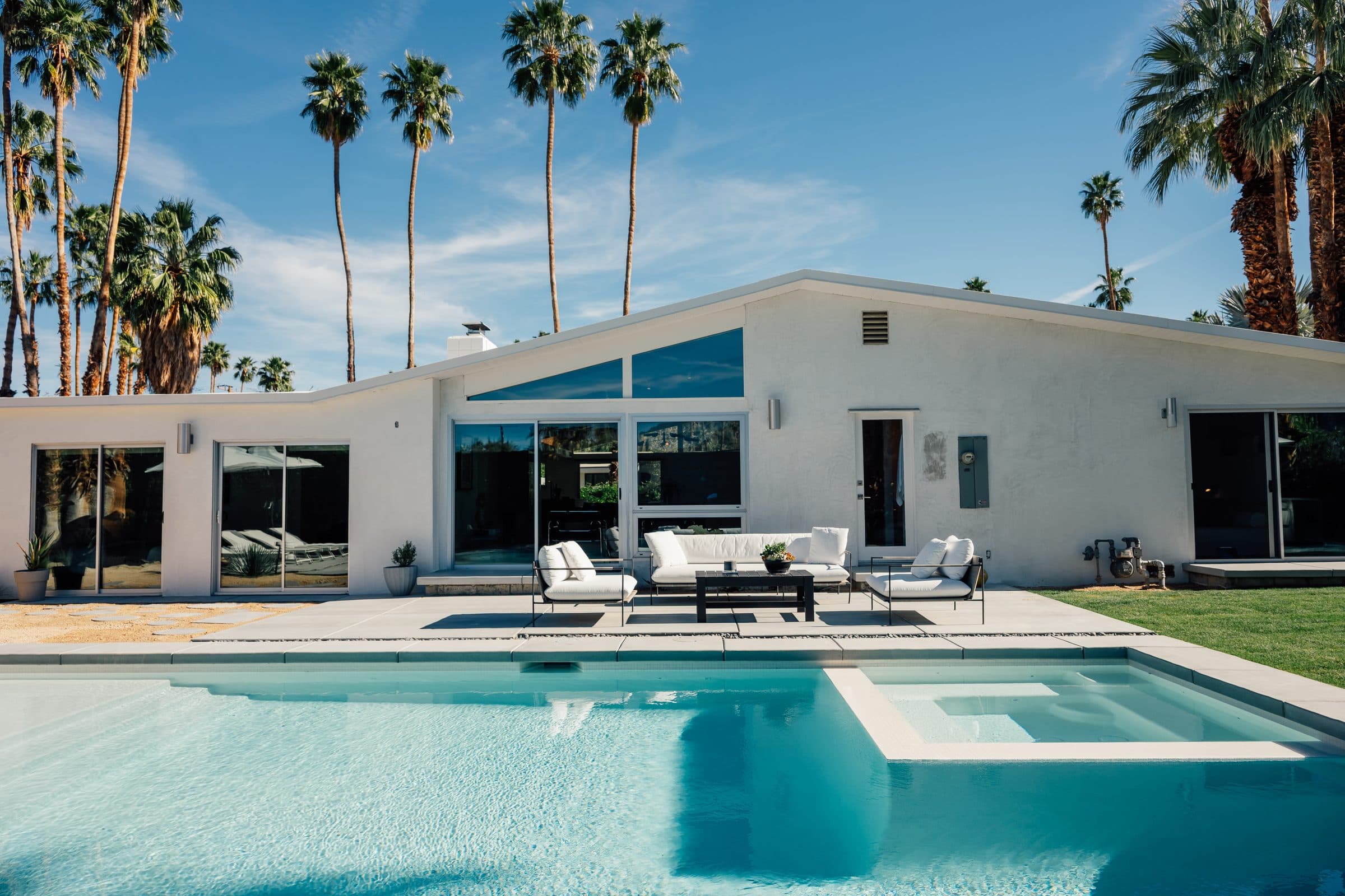 Rear elevation of the renovated 1961 Alexander home: an asymmetric gabled roofline with a triangular glass peak, full-height glass opening to the new pool and spa, and a row of tall palms behind.