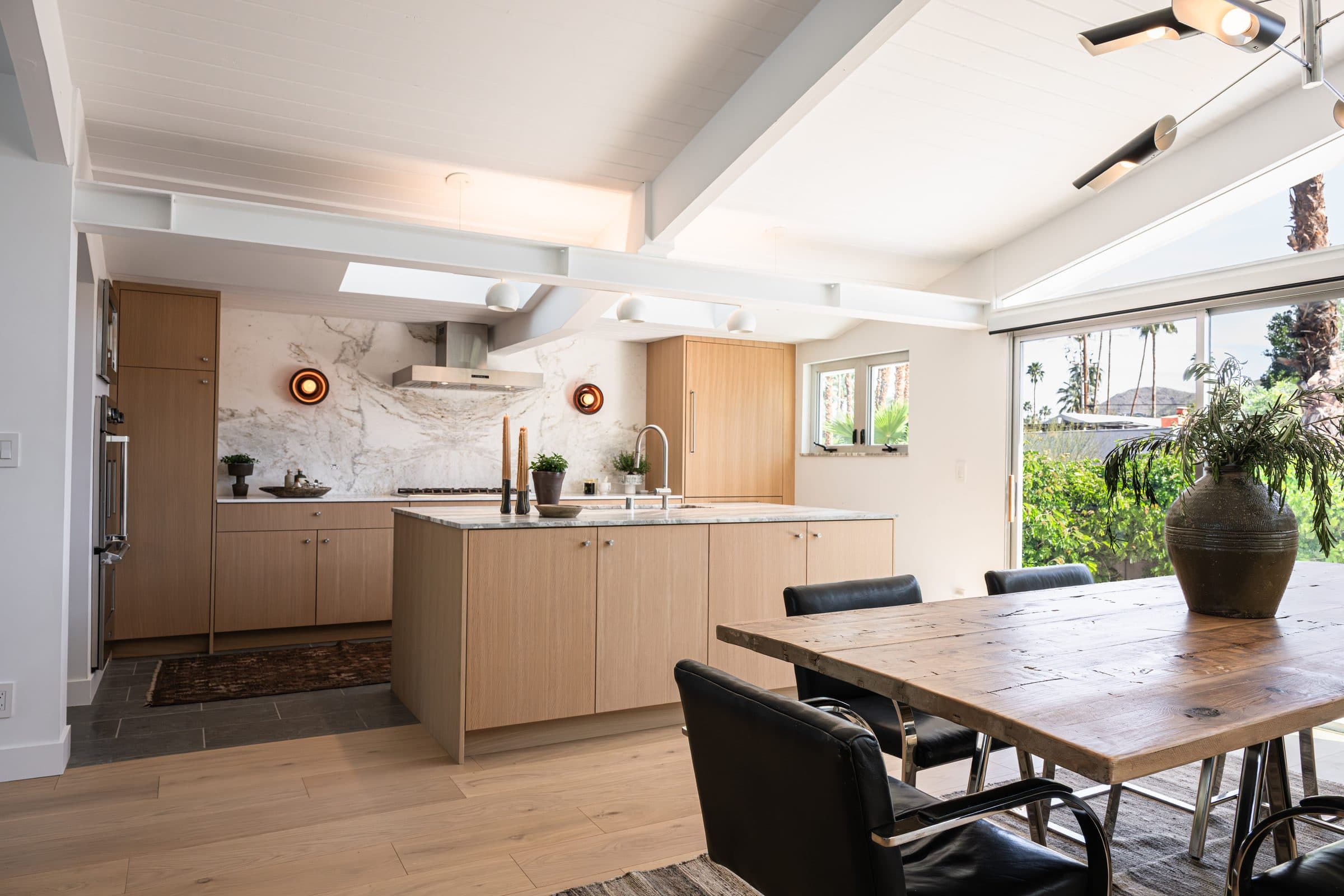 The great room from the dining table: the oak kitchen and island at center, beams overhead, and the glass wall to the backyard at right.