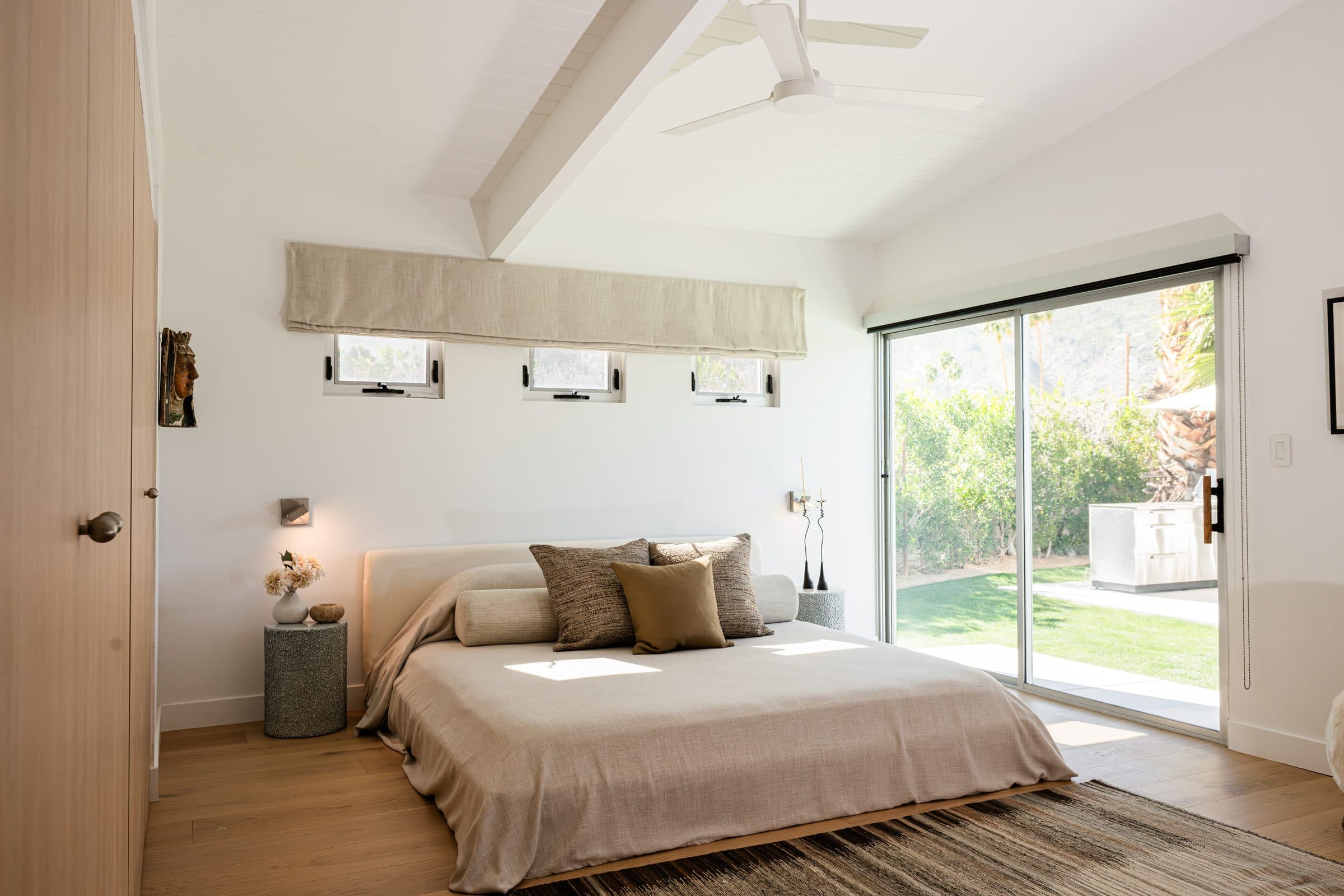 Bed beneath a vaulted white-beam ceiling, three small windows above the headboard, and a slider open to the garden.