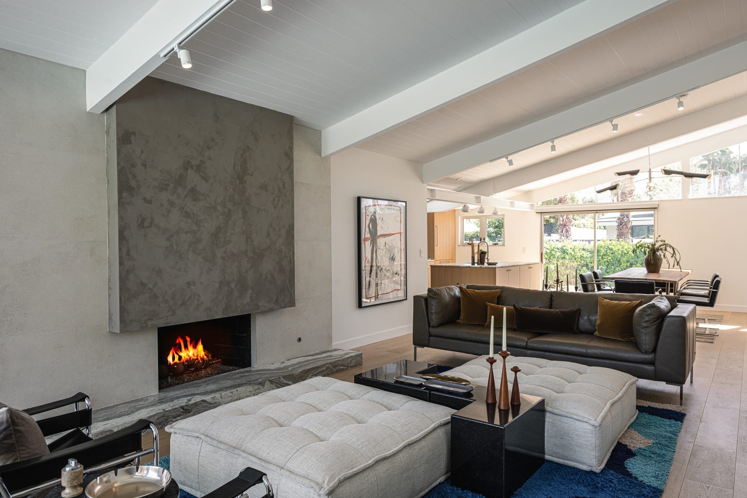 Open-plan living room under the exposed post-and-beam ceiling, looking past the seating toward the dining area and kitchen, the gray plaster fireplace lit at left.