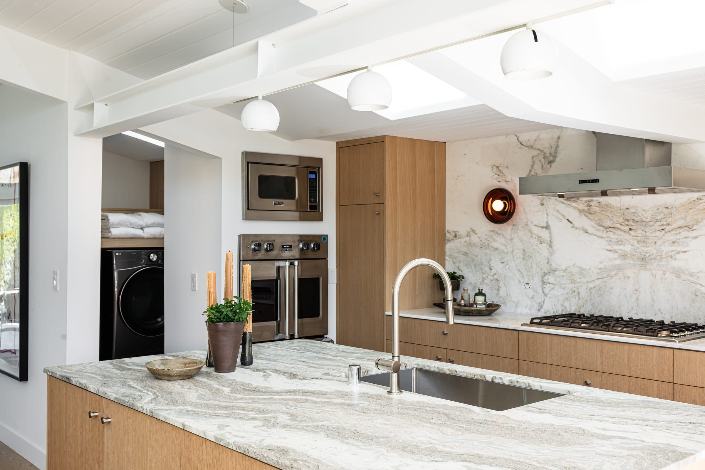 The renovated kitchen viewed across its stone-top island: bleached white oak cabinetry and a book-matched stone backsplash with a copper sconce.
