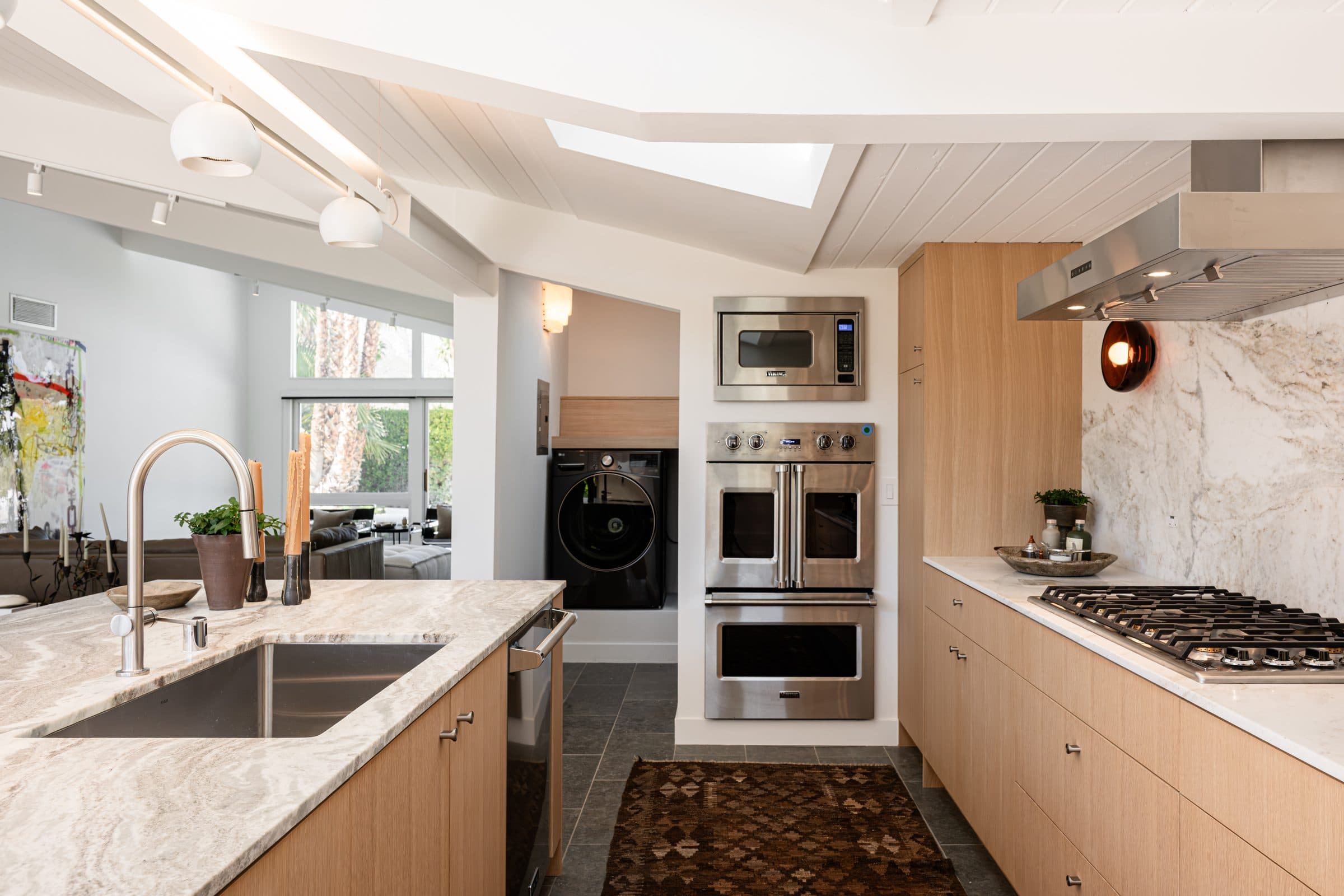 A full-slab stone backsplash behind the gas cooktop and steel hood, a copper sconce at its edge and the stacked wall ovens across the room.