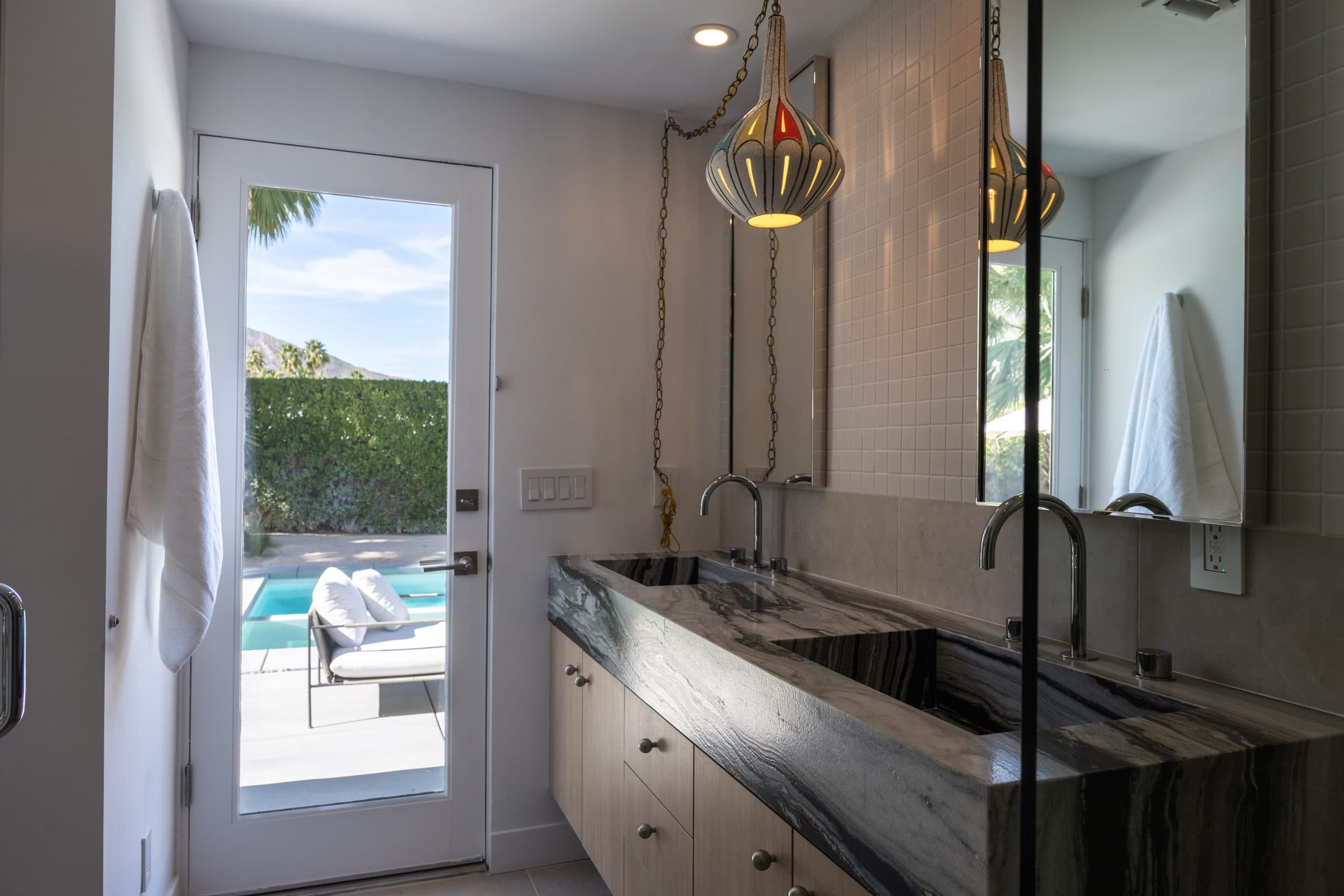 Primary bathroom opening to the pool patio: a pendant lamp on a brass chain over the double stone vanity, the San Jacinto mountains through the glass door.