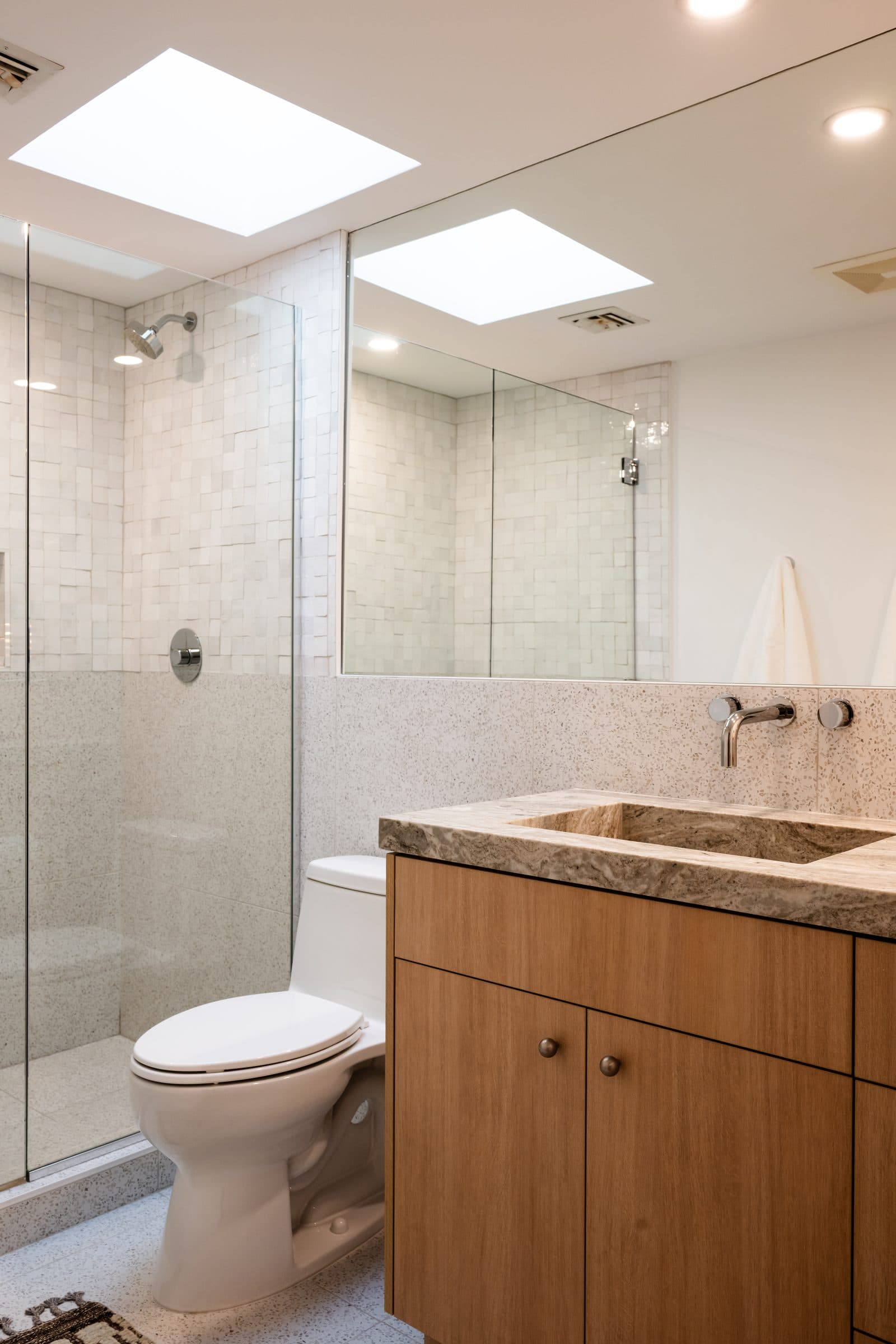 A skylit bathroom with a zellige-tile walk-in shower, terrazzo surround, a carved travertine sink in an oak vanity, and wall-mounted chrome fixtures.