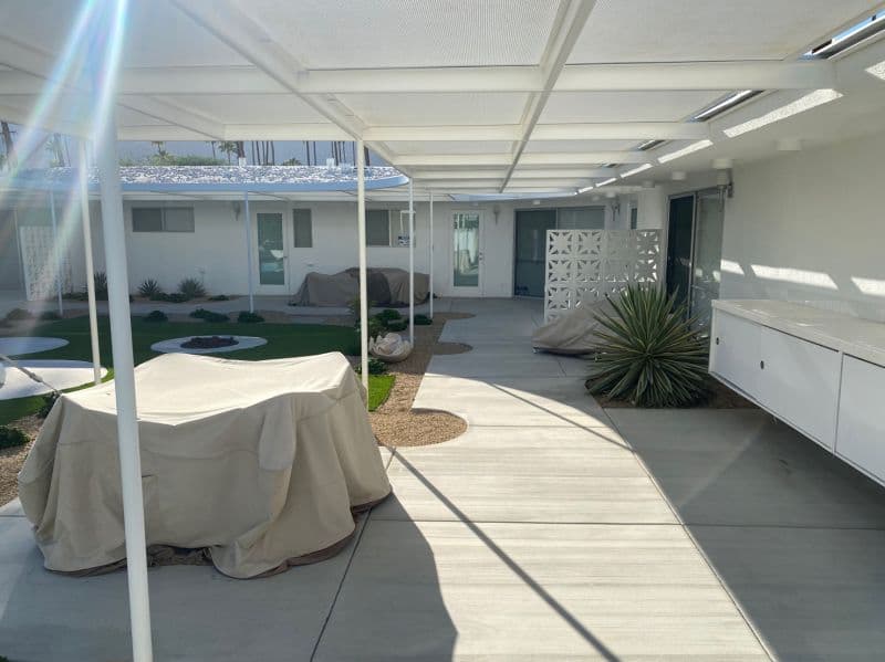 Courtyard under a white steel trellis: a breeze-block screen, an outdoor cabinet run, and furniture under covers between concrete and turf.