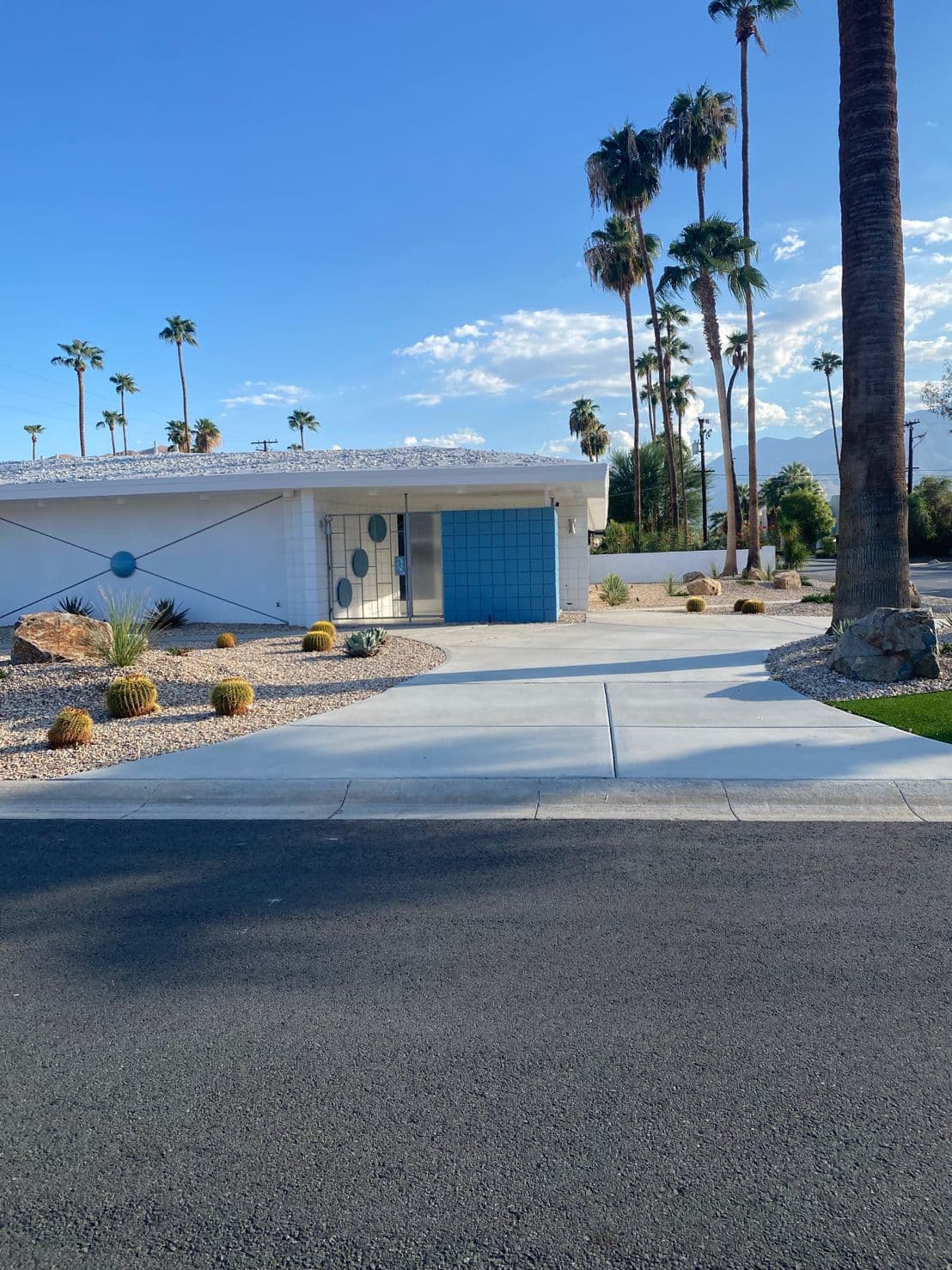 Street view of the renovated home: white walls and a rock roof, a blue-tiled entry behind a porthole-dotted gate, barrel cacti and boulders along the drive, palms behind.