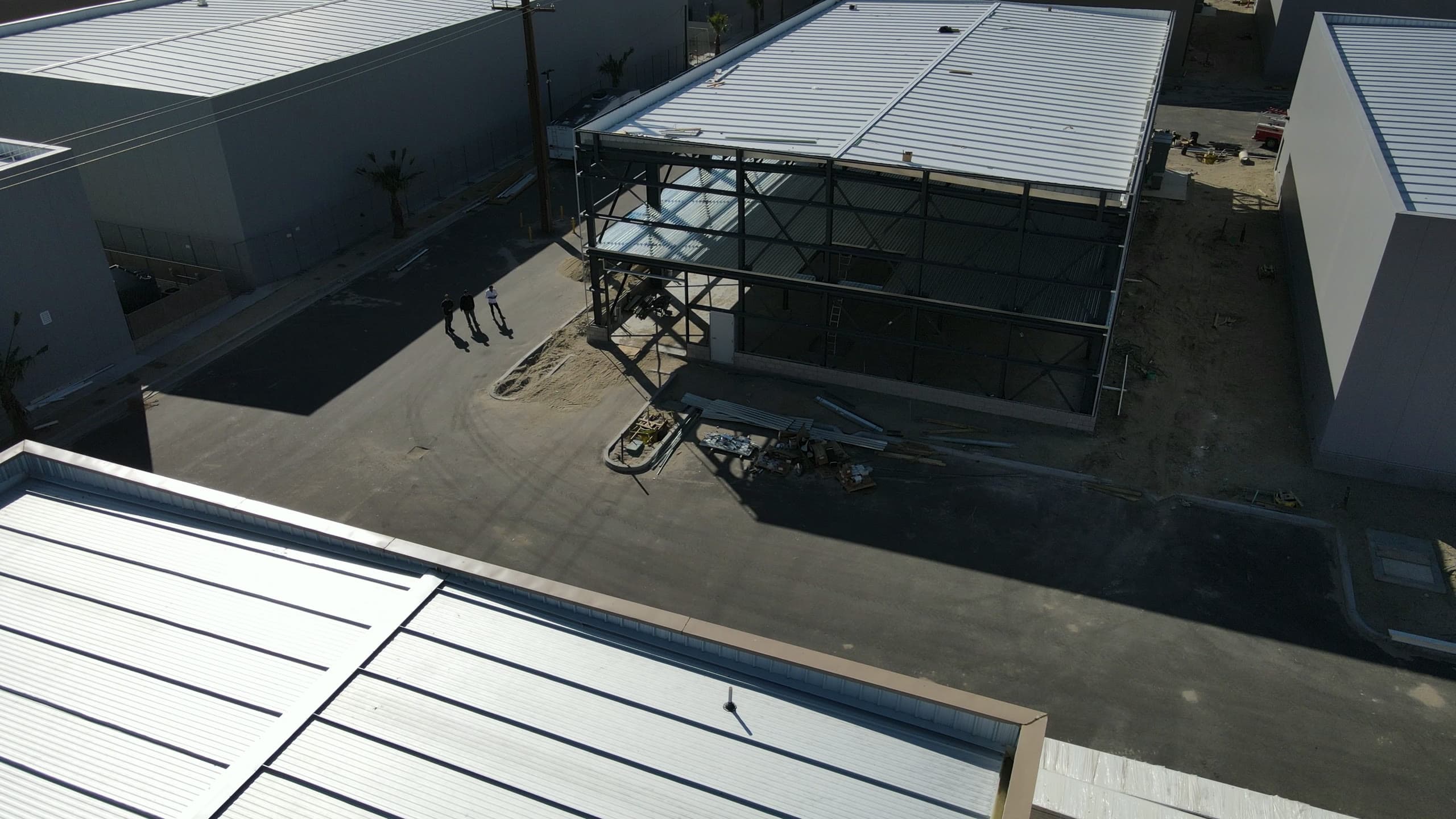 Two-story steel building frame with white roof panels over the far bays, three people walking the paved drive below, and finished metal buildings on either side.