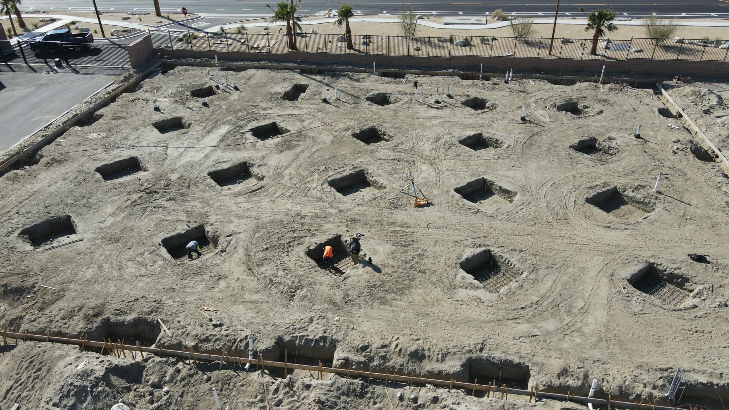 Grid of square excavated footings across a sand pad, workers tying rebar mats in two of them, white plumbing stubs in rows, and a block wall with iron fencing and palms along the street.