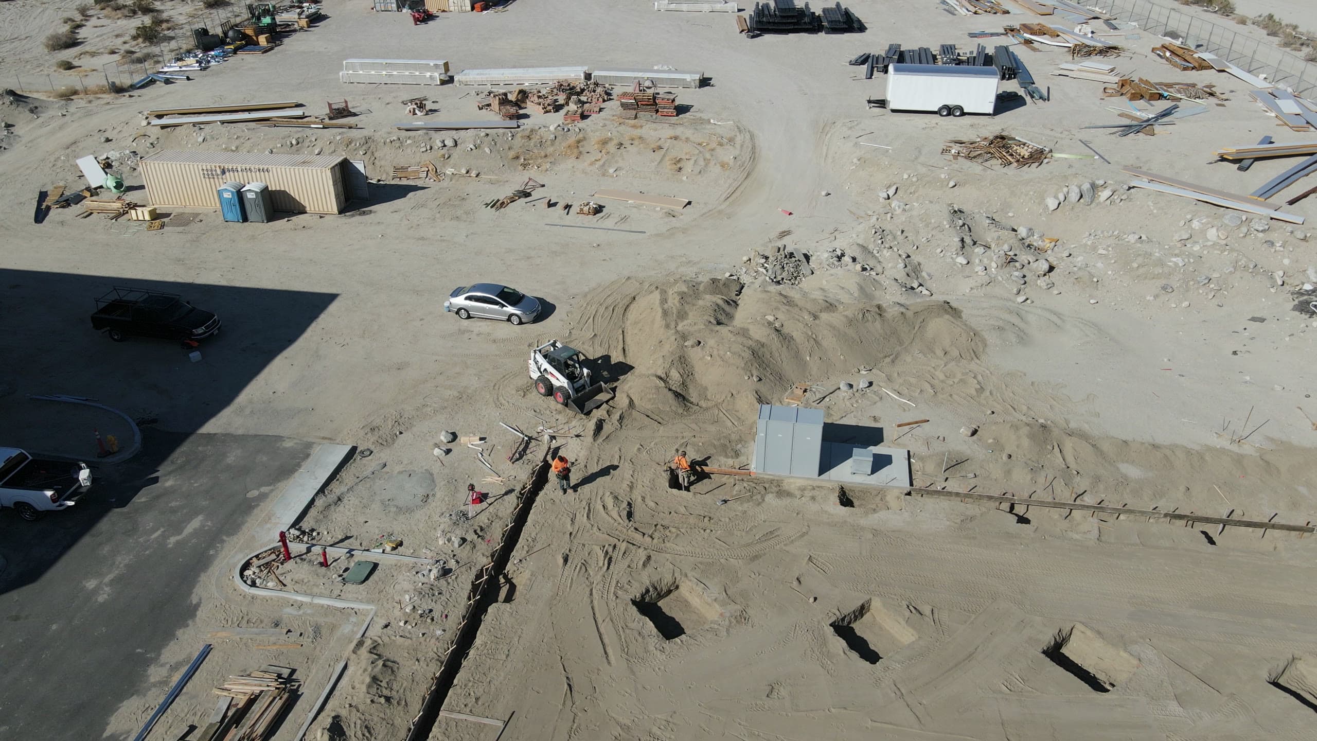 Laydown yard between pads: storage container, portable toilets, stacked steel and formwork, a skid steer moving sand in the yard, new curb and gutter along the lot edge, and workers along a utility trench.