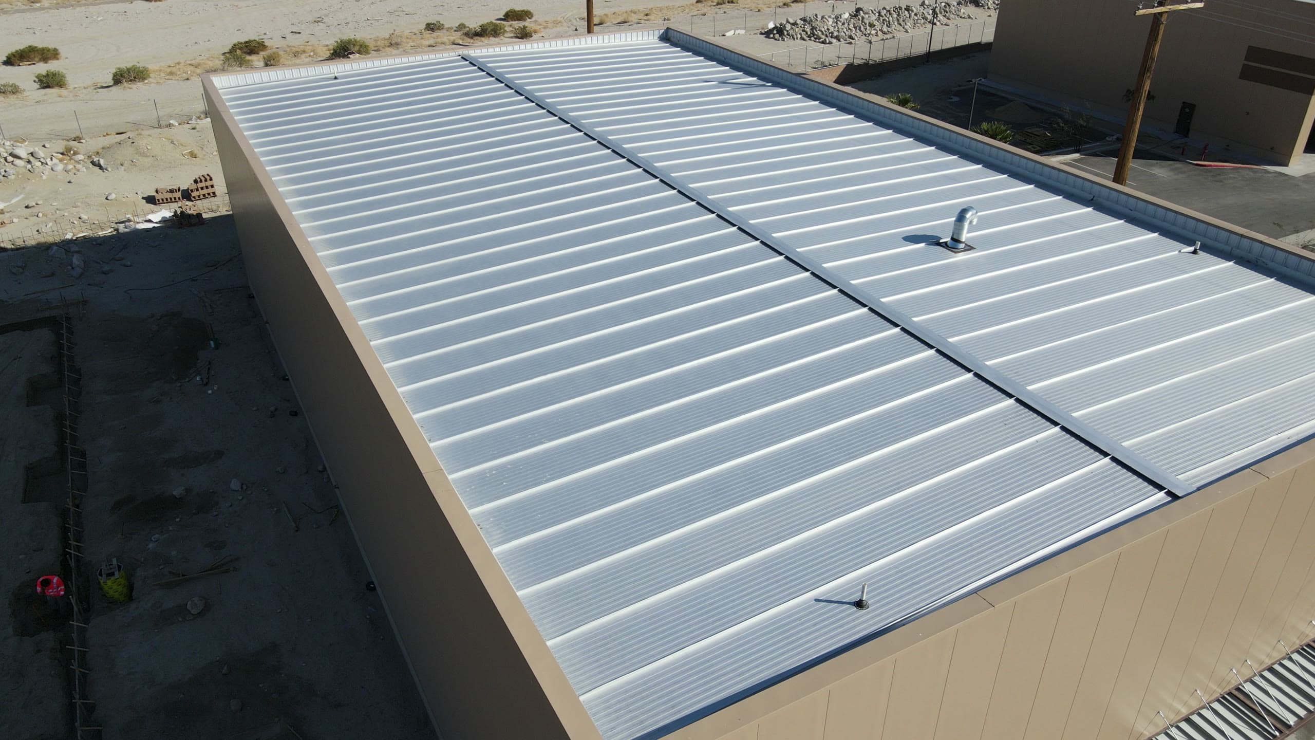 Finished white standing-seam metal roof with a plumbing vent, over tan insulated panel walls, with the neighboring building's paved yard and a utility pole beyond the parapet.