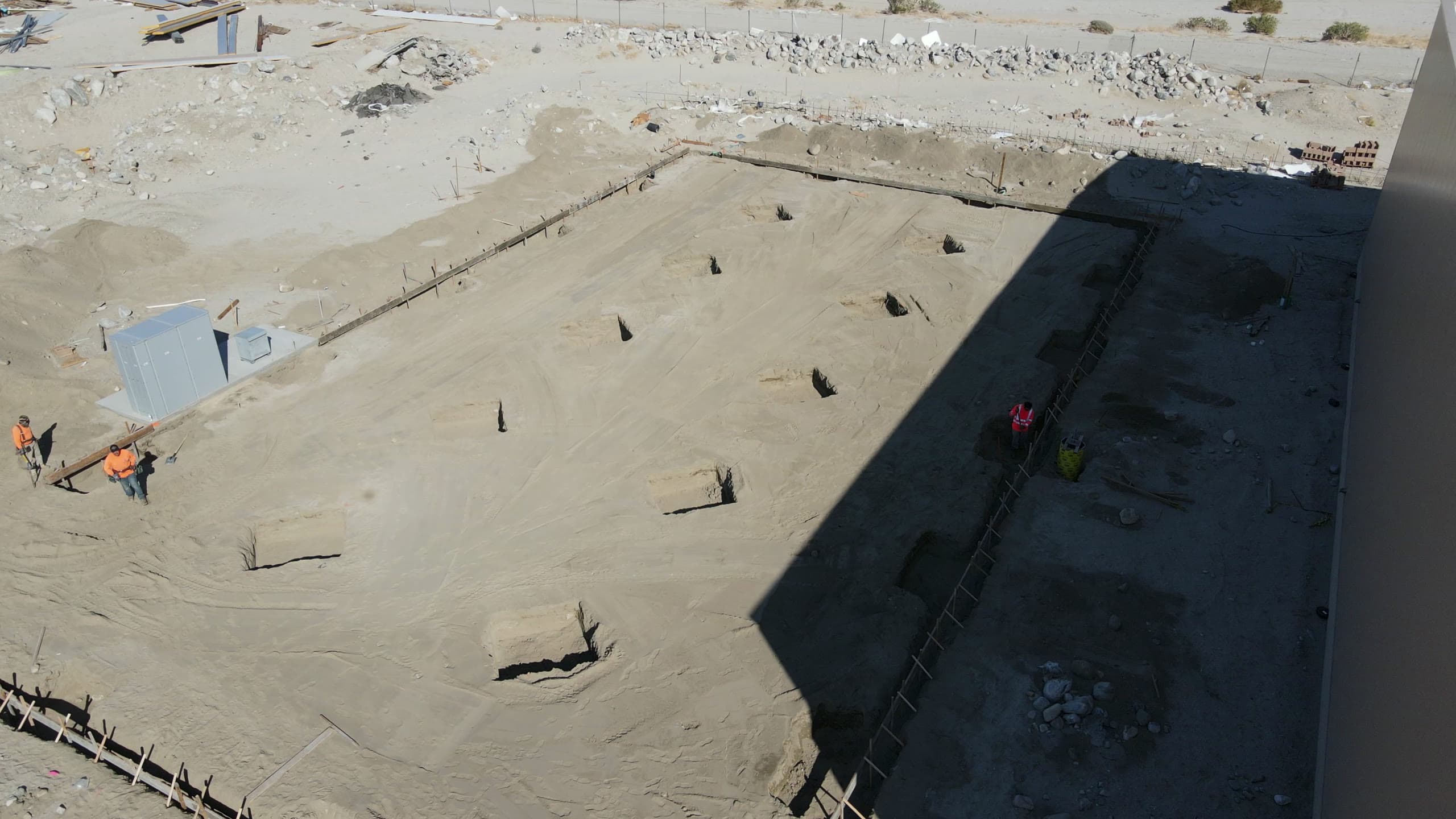 Excavation phase beside a finished building: square footings and a perimeter trench cut into the pad, workers moving material at the edge and another in the shadowed trench line.