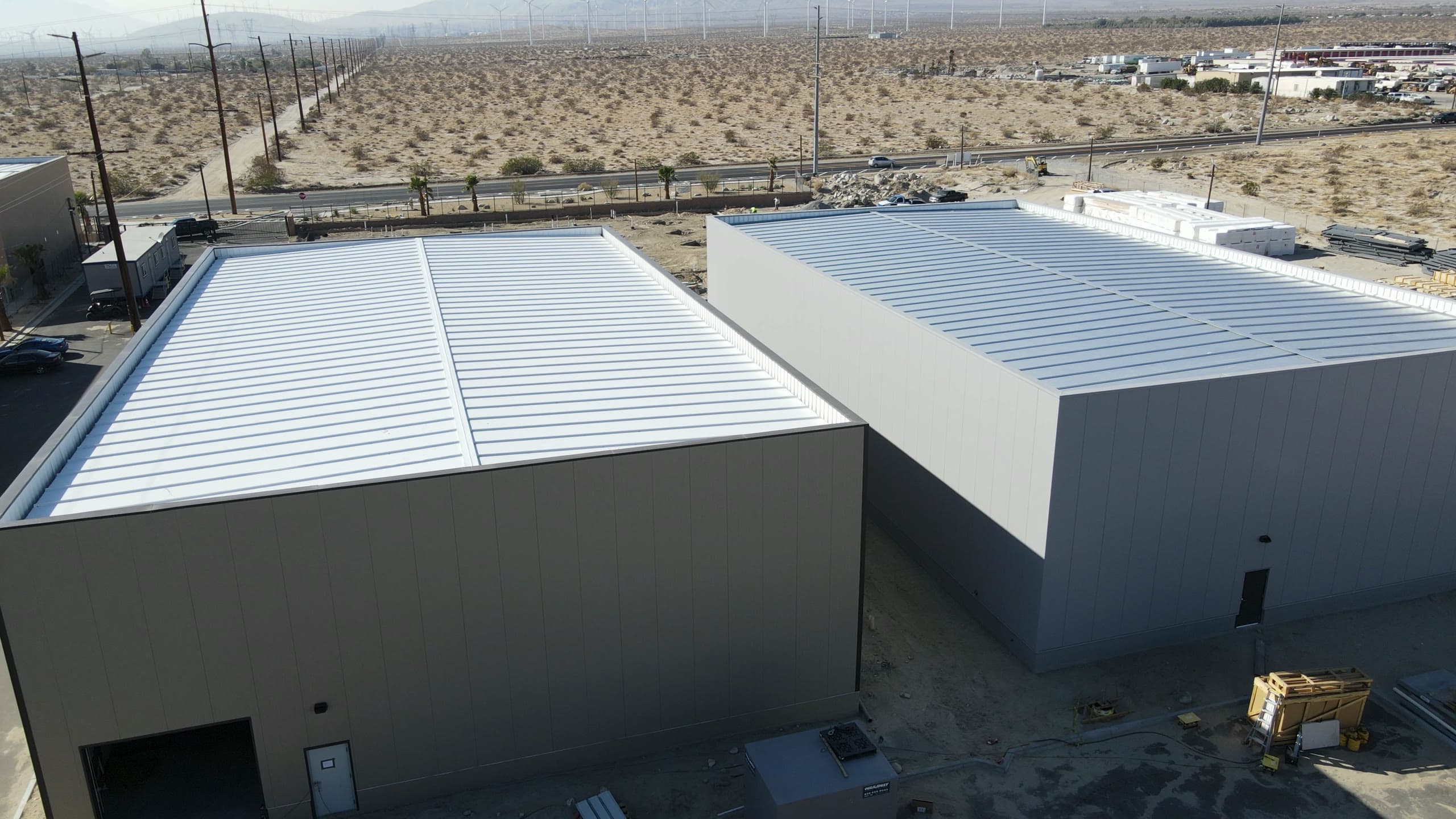 Two finished buildings in gray insulated metal panel with white standing-seam roofs, insulation bundles staged behind them and open desert across the road.