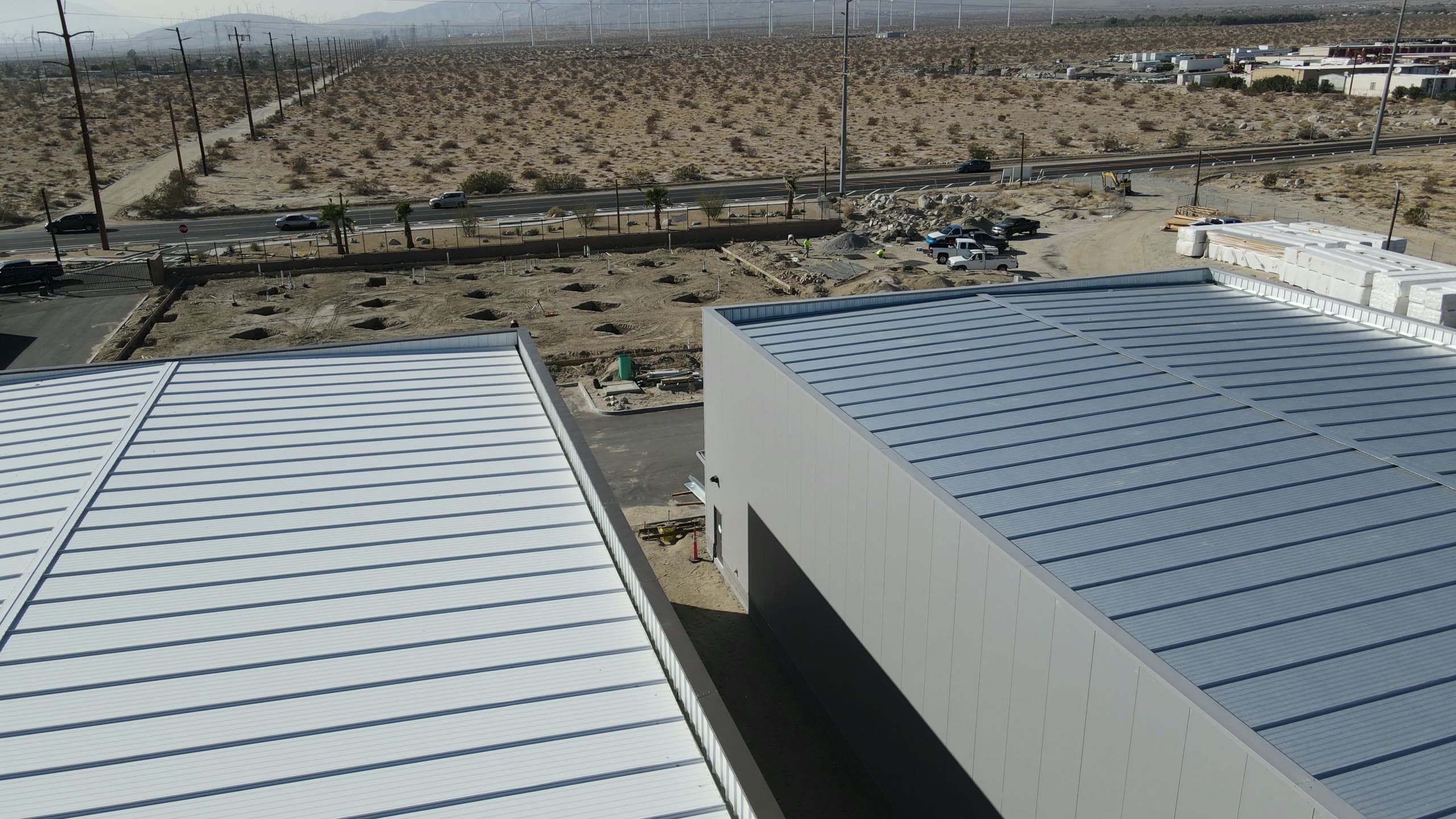 View across two finished roofs to a field of excavated footings, crew trucks along the drive, and wind turbines lining the valley floor beyond the highway.