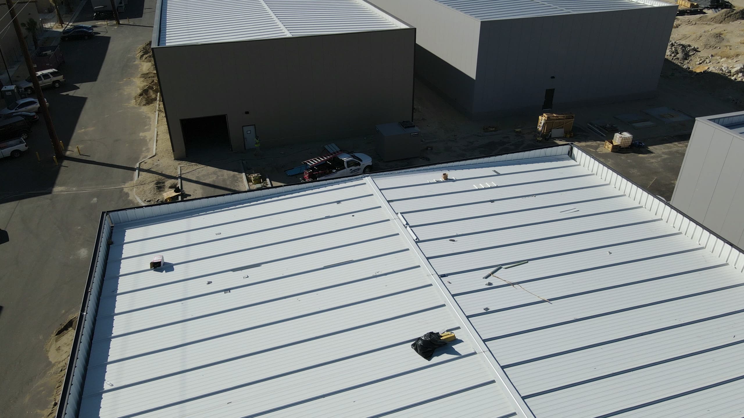 Service truck with a ladder rack parked between a finished white roof and the dark-paneled building next door, a worker in a high-visibility vest at its door and materials staged along the drive.